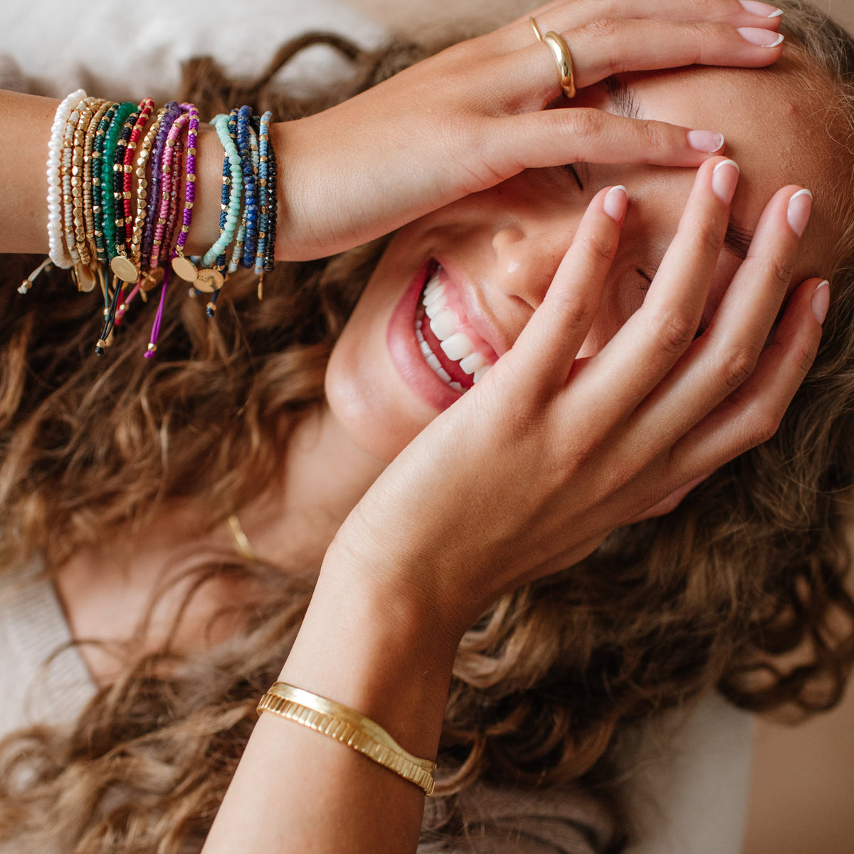 Woman with curly hair and colorful bracelets covering her face, smiling.
