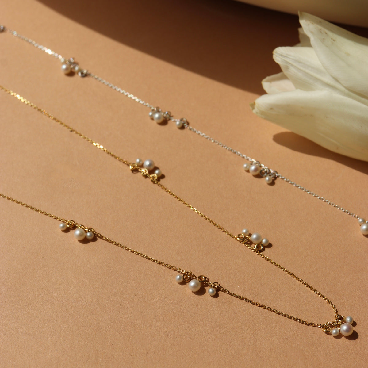 Gold necklaces with pearl drops on a beige surface with a white flower.