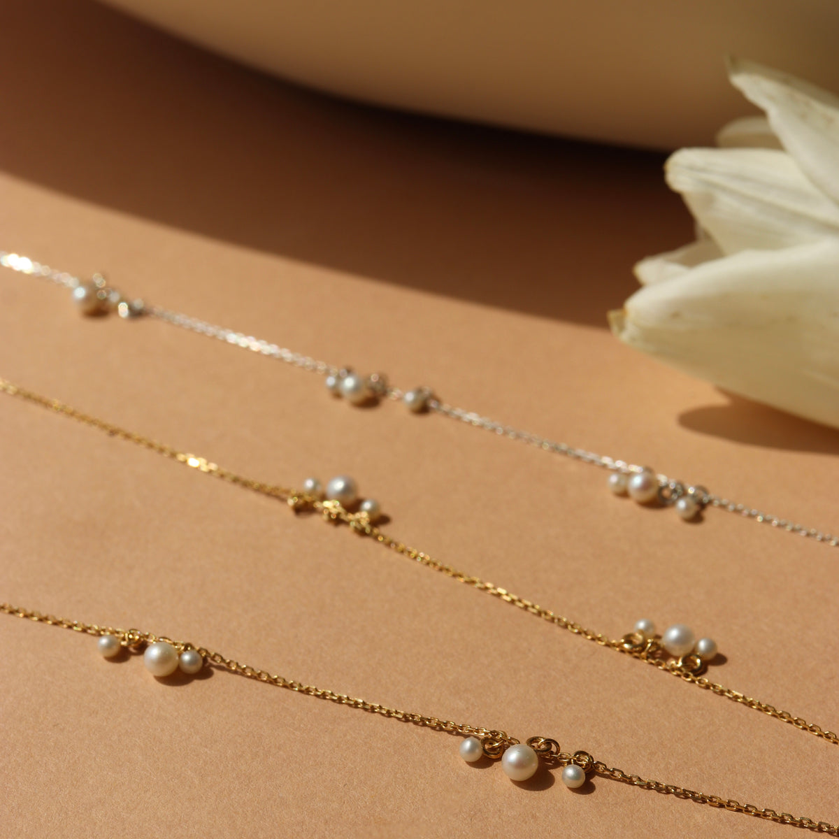 Two gold necklaces with pearls on a beige surface with a white flower.