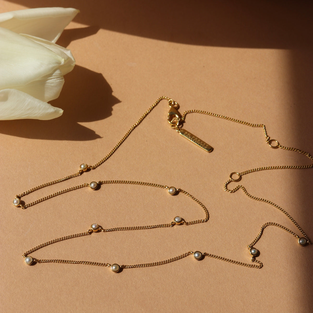Gold necklace with pearl-like beads on a beige surface with a white tulip.
